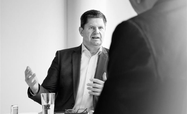 Ein Schwarz-Weiß-Foto eines Mannes in einem Anzug, der an einem Tisch sitzt und gestikulierend spricht. Vor ihm befinden sich ein Glas Wasser, eine Brille und Dokumente. Im Vordergrund ist eine weitere Person unscharf von hinten zu sehen, die offenbar an der Besprechung 