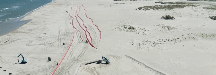 Das Foto zeigt eine Luftaufnahme vom Strand der Insel Norderney, auf dem zwei Bagger und roten Leerrohre für die Stromkabel zu sehen sind.