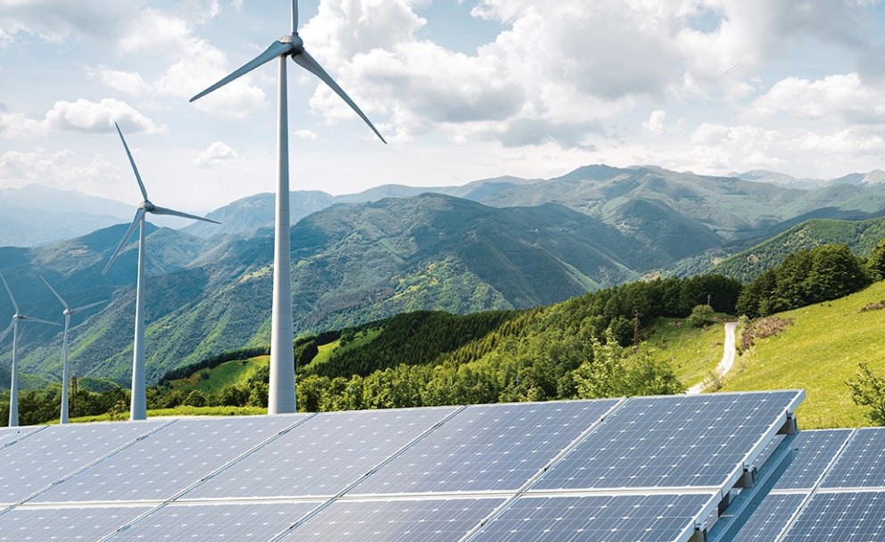 Das Bild zeigt eine weite Landschaft mit einem klaren, blauen Himmel, der von einigen weißen Wolken durchzogen ist. Im Vordergrund sind mehrere Reihen von Solarpanels zu sehen, die in einem leichten Winkel zur Sonne ausgerichtet sind. Dahinter erstrecken sich sanfte, grüne Hügel und bewaldete Gebiete, die sich bis zu einer imposanten Bergkette im Hintergrund erheben. Auf den Hügeln im Mittelgrund stehen mehrere schlanke Windräder mit ihren typischen drei Rotorblättern. Die Windräder sind in unterschiedlicher Entfernung und Größe dargestellt, was die Tiefe der Landschaft betont. Ein schmaler, heller Weg schlängelt sich einen der grünen Hänge hinauf. Die Szene vermittelt einen Eindruck von erneuerbarer Energieerzeugung in einer natürlichen und bergigen Umgebung. Das Licht deutet auf einen sonnigen Tag hin, der die Farben der Landschaft und die Reflexionen auf den Solarpanels hervorhebt.