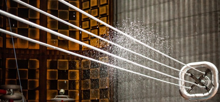 Das Bild zeigt mehrere dicke, weiße Kabel, die gespannt in einem Bogen nach oben führen. Am Ende der Kabel entspringt ein kräftiger Wasserstrahl, der in feine Tropfen zerstäubt und eine Art Fontäne bildet. Der Hintergrund ist unscharf und zeigt eine metallische Wand mit einer quadratischen Musterung. Die Szene wirkt industriell und technisch. Das Zusammenspiel von den glatten, weißen Kabeln und dem zerstäubenden Wasser schafft einen interessanten Kontrast zwischen Ruhe und Bewegung. Die Beleuchtung ist kühl und sachlich, was die technische Atmosphäre verstärkt.