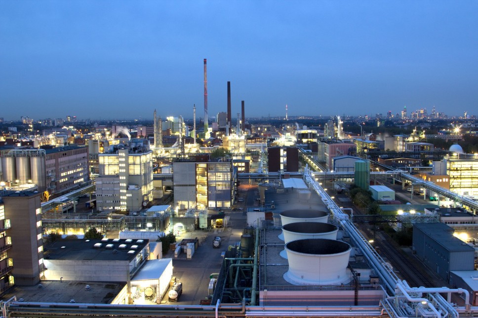 Ein großflächiges Industriegebiet bei Dämmerung mit zahlreichen beleuchteten Gebäuden, Schornsteinen und Produktionsanlagen. Im Hintergrund erstreckt sich eine Stadtlandschaft mit Hochhäusern und einem Fernsehturm.