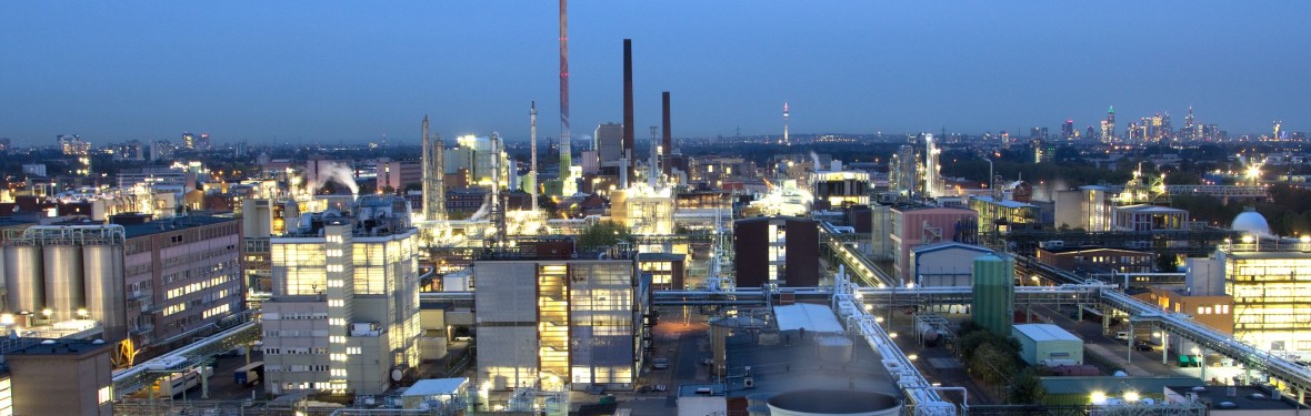 Ein großflächiges Industriegebiet bei Dämmerung mit zahlreichen beleuchteten Gebäuden, Schornsteinen und Produktionsanlagen. Im Hintergrund erstreckt sich eine Stadtlandschaft mit Hochhäusern und einem Fernsehturm.