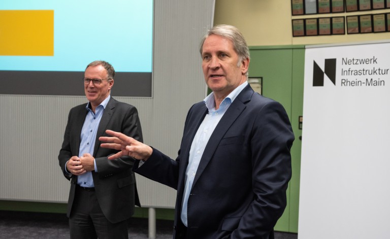 Zwei Männer in Anzügen stehen in einem Konferenzraum. Der Mann im Vordergrund gestikuliert, während der andere lächelnd im Hintergrund steht. Rechts befindet sich ein Banner mit der Aufschrift „Netzwerk Infrastruktur Rhein-Main“. Eine Leinwand mit einer Präsentationsfolie ist ebenfalls sichtbar.