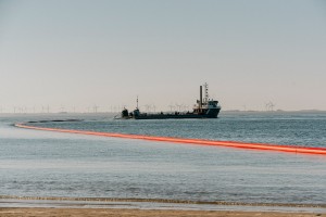 Das Bild zeigt ein Schiff, das in einem Gewässer vor Anker liegt. Hinter dem Schiff erstreckt sich eine lange, orangefarbene Rohrleitung über das Wasser bis zum Ufer. Im Hintergrund sind Windräder zu erkennen.