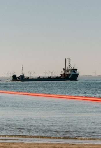 Das Bild zeigt ein Schiff, das in einem Gewässer vor Anker liegt. Hinter dem Schiff erstreckt sich eine lange, orangefarbene Rohrleitung über das Wasser bis zum Ufer. Im Hintergrund sind Windräder zu erkennen. 