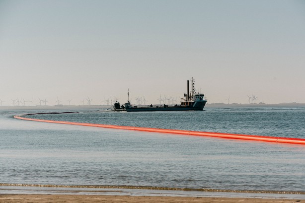 Das Bild zeigt ein Schiff, das in einem Gewässer vor Anker liegt. Hinter dem Schiff erstreckt sich eine lange, orangefarbene Rohrleitung über das Wasser bis zum Ufer. Im Hintergrund sind Windräder zu erkennen.
