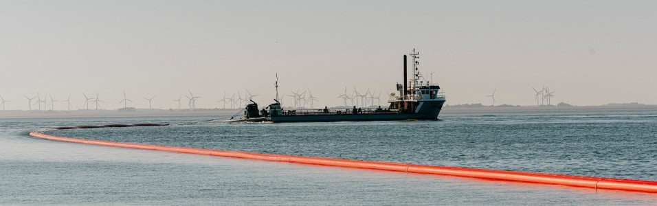 Das Bild zeigt ein Schiff, das in einem Gewässer vor Anker liegt. Hinter dem Schiff erstreckt sich eine lange, orangefarbene Rohrleitung über das Wasser bis zum Ufer. Im Hintergrund sind Windräder zu erkennen. 