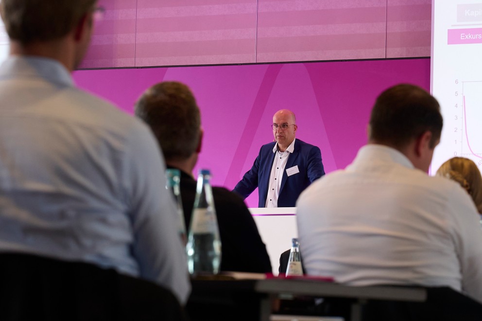 Ein Konferenzraum mit mehreren Personen, die einem Vortrag zuhören. Im Vordergrund sind unscharf die Rückenansichten der Teilnehmenden zu sehen, die an Tischen mit Wasserflaschen und Notizmaterial sitzen. Im Hintergrund steht ein Mann im Anzug an einem Rednerpult und hält einen Vortrag. Hinter ihm ist eine große Präsentationsleinwand mit einer Folie zu sehen, die den Titel "Netzentgeltentwicklung in fünf Faktoren" trägt. Die Beleuchtung des Raums ist modern, mit deckenmontierten Lampen und einem hell-lila Lichtakzent auf der Bühne.