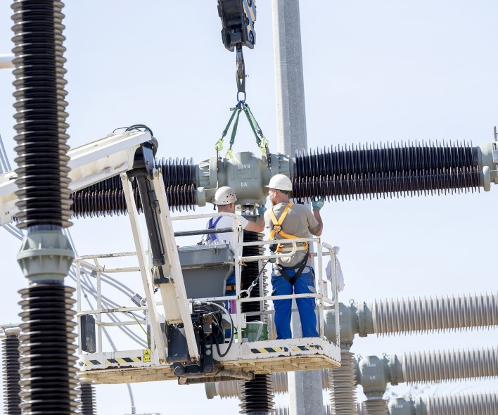 Das Bild zeigt zwei Menschen, die auf einer Hebebühne in einer Umspannanlage  arbeiten. Sie sind mit Schutzhelmen und Sicherheitsgurten ausgestattet, um sich vor den Gefahren des Stroms zu schützen. 