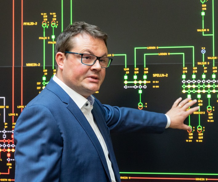 Ein Mann, Dr. Christoph Schneiders, steht in Geschäftskkleidung vor einem schwarzen Monitor mit bunt leuchtenden Schaltkreisen. Er deutet sprechend mit seiner linken Hand auf einen Bereich des Monitors.