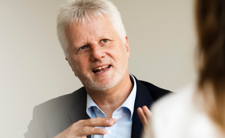 Gesprächssituation: Ein Mann mit weißem Haar und kurzem Bart, gekleidet in einem Anzug mit hellblauem Hemd, spricht gestikulierend. Er sitzt an einem Tisch vor einem neutralen, hellen Hintergrund. Im Vordergrund ist unscharf die Schulter einer anderen Person sichtbar, die ihm gegenüber sitzt. Der Mann wirkt engagiert und konzentriert.
