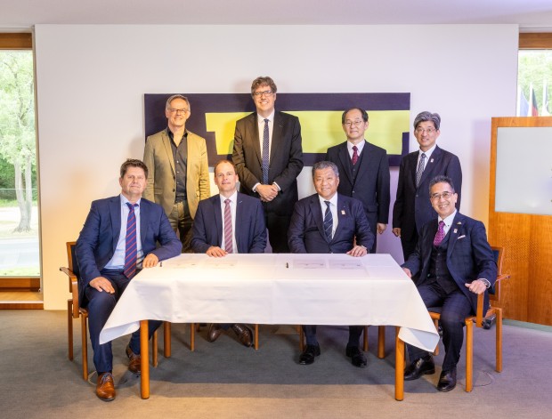 Acht Personen in zwei Reihen posieren für ein Gruppenfoto. Die vordere Reihe zeigt vier sitzende Männer an einem Tisch mit weißer Tischdecke, auf dem offenbar ein Vertrag oder Dokument liegt. Dahinter stehen vier weitere Männer. Die Teilnehmer sind in formeller Kleidung, und im Hintergrund hängt ein Kunstwerk mit gelben und dunklen Formen an der Wand. Die Umgebung ist ein heller Konferenzraum mit Blick ins Grüne.