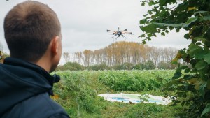 Blick über die Schulter eines Mannes in dunkler praktischer Kleidung. Der Mann blickt auf ein Feld. Über dem Feld schwebt eine Drohne.
