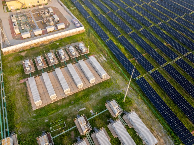Luftaufnahme eines Solarparks mit Reihen von Solarmodulen und angrenzenden weißen Batteriecontainern auf Grasfläche