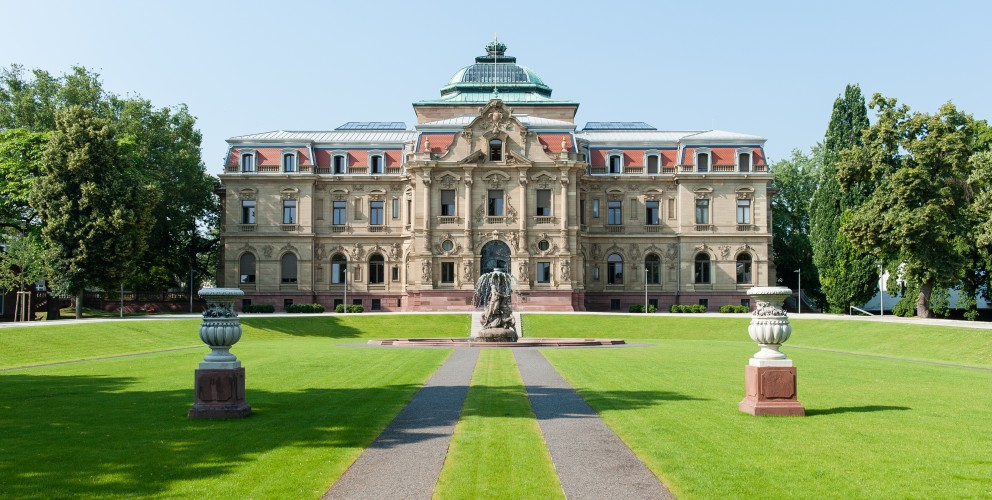 Palais des Bundesverfassungsgerichts vor blauem Himmer. Im Vordergrund eine gepflegte Parkanlage.