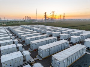 Zu sehen sind mehrere, linear angeordnete große Container mit Batteriespeichern. Dahinter sind Strommasten und Leitungen zu erkennen. 