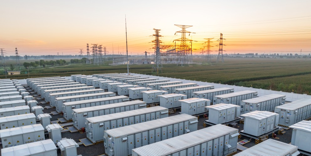 Zu sehen sind mehrere, linear angeordnete große Container mit Batteriespeichern. Dahinter sind Strommasten und Leitungen zu erkennen. 