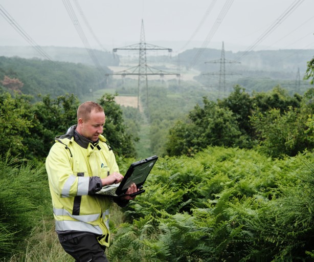 Ein Mann in einer gelben Warnjacke steht in einer grünen, bewaldeten Landschaft und schaut auf einen Laptop, den er in den Händen hält. Im Hintergrund sind mehrere große Strommasten und Stromleitungen unter einem hellgrauen Himmel zu sehen. Die Vegetation besteht hauptsächlich aus niedrigen Büschen und Bäumen.