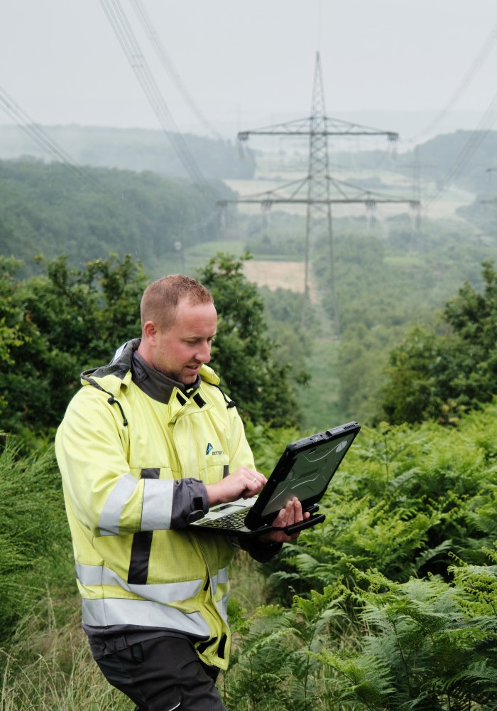 Ein Mann in einer gelben Warnjacke steht in einer grünen, bewaldeten Landschaft und schaut auf einen Laptop, den er in den Händen hält. Im Hintergrund sind mehrere große Strommasten und Stromleitungen unter einem hellgrauen Himmel zu sehen. Die Vegetation besteht hauptsächlich aus niedrigen Büschen und Bäumen.