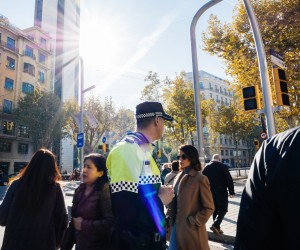 Im Vordergund ein spanischer Poluizist mit Blick auf die mehrspurige Straße in großstädtischem Stadtbild. Hinter ihm queren Passant:innen die Straße. Die Ampeln sind ausgefallen und zeigen keine Signale. Die Sonne scheint. 