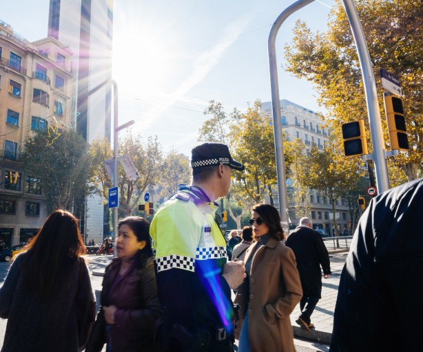 Im Vordergund ein spanischer Poluizist mit Blick auf die mehrspurige Straße in großstädtischem Stadtbild. Hinter ihm queren Passant:innen die Straße. Die Ampeln sind ausgefallen und zeigen keine Signale. Die Sonne scheint. 