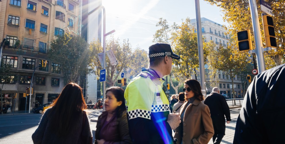 Im Vordergund ein spanischer Poluizist mit Blick auf die mehrspurige Straße in großstädtischem Stadtbild. Hinter ihm queren Passant:innen die Straße. Die Ampeln sind ausgefallen und zeigen keine Signale. Die Sonne scheint.