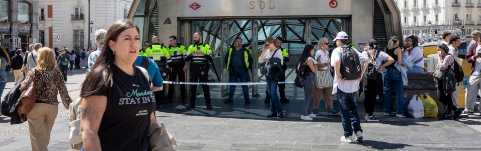 Polizisten in gelb-schwarzen Uniformen stehen vor dem Eingang der U-Bahn-Station Sol in einer belebten Straße mit Passanten und Gebäuden im Hintergrund.