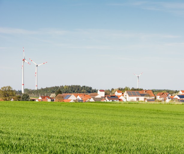 Ein grünes Feld im Vordergrund, im Hintergrund ein Dorf. Auf einigen Dächern befinden sich PV-Anlagen, dahinter Windräder. Die Sonne scheint, der Himmel ist wolkenfrei. 