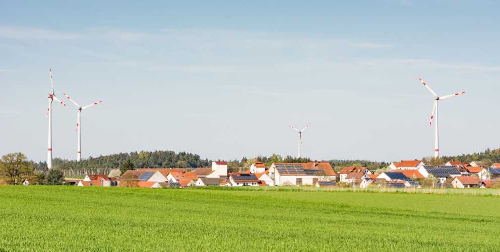 Ein grünes Feld im Vordergrund, im Hintergrund ein Dorf. Auf einigen Dächern befinden sich PV-Anlagen, dahinter Windräder. Die Sonne scheint, der Himmel ist wolkenfrei. 