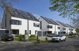 Vier moderne weiße Reihenhäuser mit jeweils zwei Stockwerken und einem Dachstuhl. Auf jedem Dach befinden sich PV-Anlagen. Vor dern Häusern kleine aufgeräumte Vorgärten und vereinzelte Pkw. Die Sonne scheint, der Himmel ist wolkenlos.