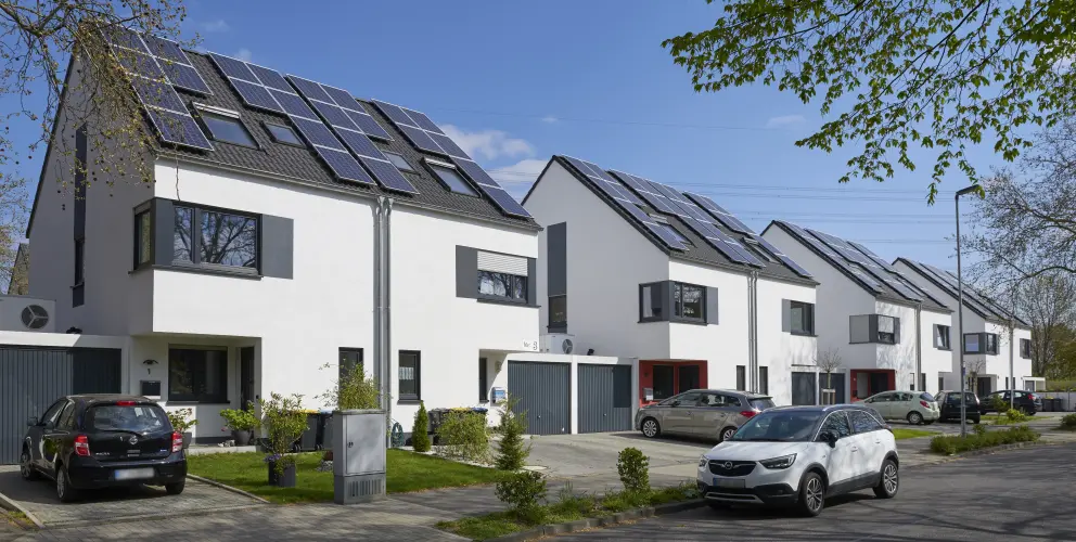 Vier moderne weiße Reihenhäuser mit jeweils zwei Stockwerken und einem Dachstuhl. Auf jedem Dach befinden sich PV-Anlagen. Vor dern Häusern kleine aufgeräumte Vorgärten und vereinzelte Pkw. Die Sonne scheint, der Himmel ist wolkenlos.