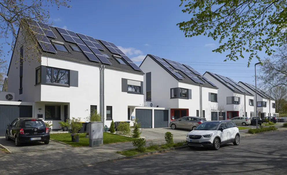 Vier moderne weiße Reihenhäuser mit jeweils zwei Stockwerken und einem Dachstuhl. Auf jedem Dach befinden sich PV-Anlagen. Vor dern Häusern kleine aufgeräumte Vorgärten und vereinzelte Pkw. Die Sonne scheint, der Himmel ist wolkenlos.