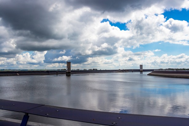 Ein großes Wasserreservoir, umgeben von einem massiven Betondamm. Zwei rechteckige Türme ragen aus dem ruhigen Wasser. Der Himmel ist bewölkt mit blauen Flecken.