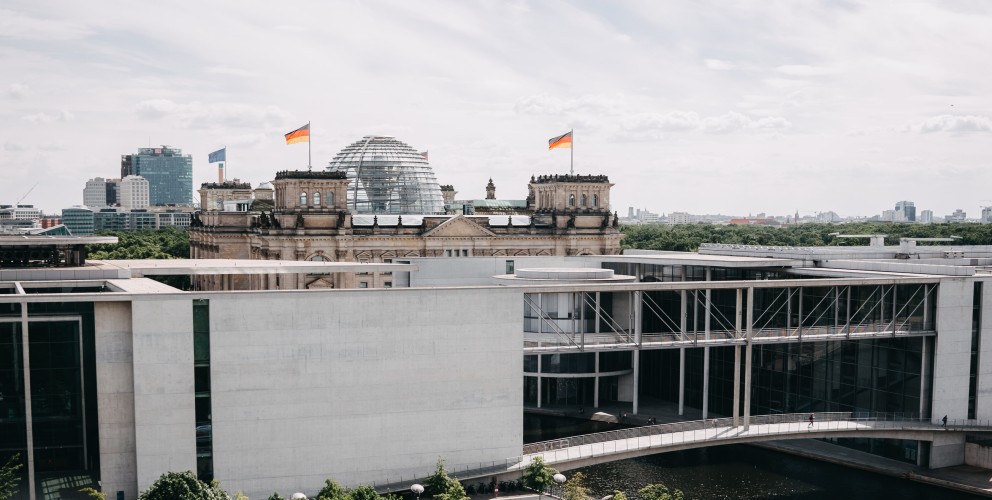 Kanzleramt bei grauem Wetter. Im Vordergrund Bäume und die Spree, im Hintergrund der Reichstag.