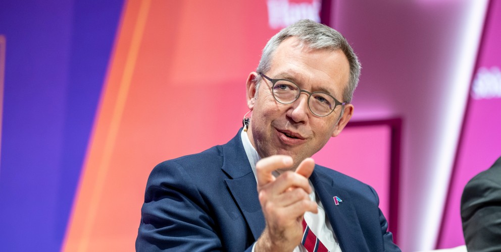 Das Bild zeigt Herrn Müller, den CEO von Amprion, in einem professionellen Umfeld. Er trägt eine Brille mit dünnem Metallrahmen, einen dunkelblauen Anzug, ein weißes Hemd und eine dezente Krawatte. Auf dem Revers seines Anzugs befindet sich das Amprion-Logo als kleines Abzeichen. Herr Müller lächelt leicht und gestikuliert mit der rechten Hand, als würde er etwas erklären. Am Ohr ist eine Mikrofonhalterung sichtbar, was darauf hinweist, dass er gerade spricht. Der Hintergrund ist in dynamischen Farben gehalten – Lila, Pink und Orange –, was auf eine moderne Veranstaltung oder ein Panel hinweist.
