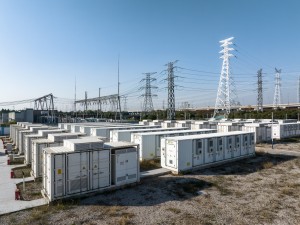 Zu sehen sind mehrere, linear angeordnete große Container mit Batteriespeichern. Dahinter sind Strommasten und Leitungen zu erkennen. 