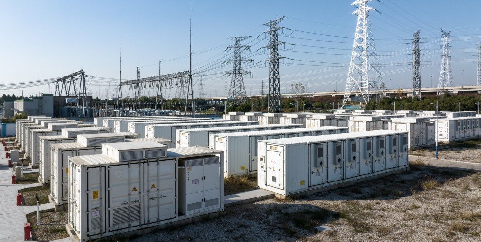 Zu sehen sind mehrere, linear angeordnete große Container mit Batteriespeichern. Dahinter sind Strommasten und Leitungen zu erkennen. 