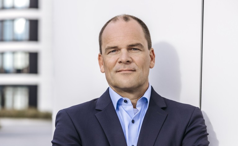 Herr Neumann lehnt mit verschränkten Armen an einer einer Faassade eines Bürogebäudes. Er trägt Geschäftskleidung und blickt direkt in die Kamera.