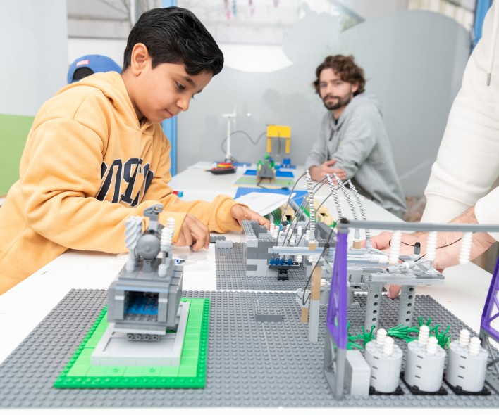 Junge in gelber Kapuzenjacke baut an einem Modell aus Legosteinen auf einem Tisch mit weiteren Modellen und Bauteilen