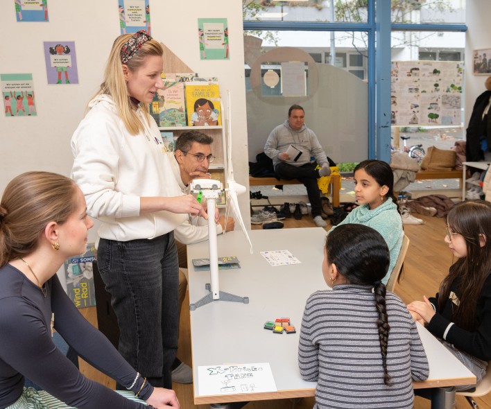 Mehrere Kinder und eine erwachsene Person stehen und sitzen an einem Tisch in einem Klassenraum mit Lernmaterialien und Postern an den Wänden