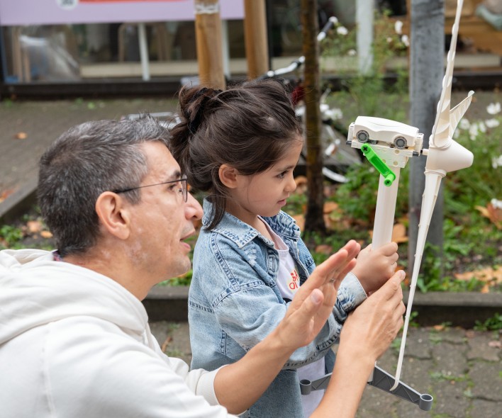 Kind hält ein kleines Windradmodell an einem Stab und wird von einer Person im weißen Kapuzenpullover unterstützt