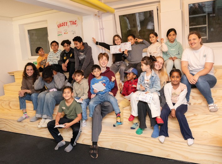 Gruppe von Kindern und Jugendlichen sitzt auf einer hölzernen Tribüne in einem Innenraum.