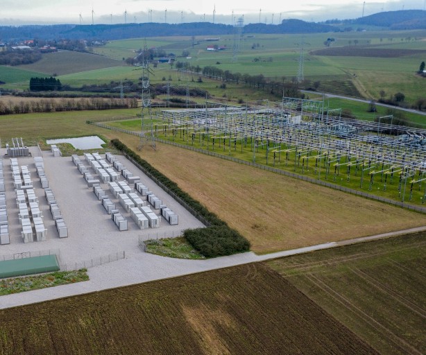 Die Luftaufnahme zeigt aus einer erhöhten Perspektive ein weitläufiges Umspannwerk (rechts) und einen direkt angrenzenden Batteriespeicher-Park (links) in einer ländlichen Umgebung.
Batteriespeicher (links): Auf einer rechteckigen, mit hellem Schotter befestigten Fläche sind über 30 große, weiße Container in zwei parallelen Doppelreihen angeordnet. Die Anlage ist von einem Schutzzaun und einer niedrigen, grünen Hecke umgeben. Am unteren Rand der Fläche befindet sich ein flaches, grünes Technikgebäude.
Umspannwerk (rechts): Direkt daneben befindet sich eine klassische Freiluft-Schaltanlage. Sie besteht aus einem dichten Netz von hohen, grauen Stahlgittermasten, Isolatorenketten und horizontal verlaufenden Stromschienen über einer Rasenfläche.
Umgebung: Die Anlagen sind in eine hügelige Landschaft mit weiten, grünen und braunen Feldern eingebettet. Im Hintergrund am Horizont sind zahlreiche Windkraftanlagen zu erkennen, was den Bezug zur Erzeugung und Speicherung erneuerbarer Energien unterstreicht. Der Himmel ist hellgrau und bedeckt.