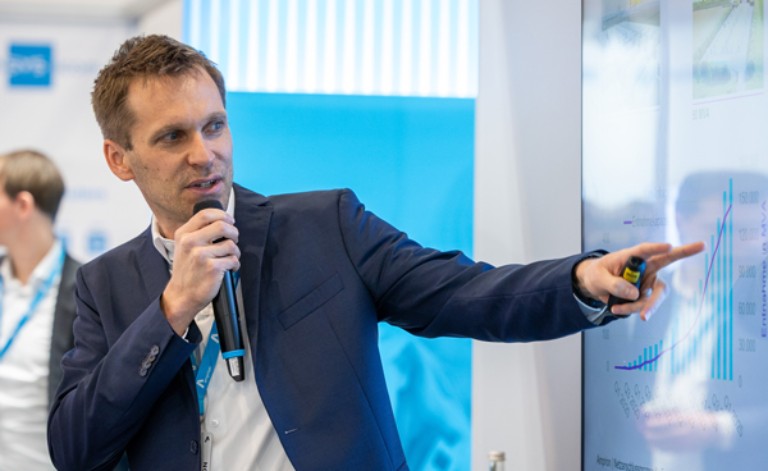 Das Foto zeigt einen Mann mittleren Alters während eines Vortrags auf einer Messe oder Fachkonferenz.
Person und Aktion: Der Mann ist in einer dynamischen Halbprofil-Ansicht von der Seite zu sehen. Er hält ein schwarzes Funkmikrofon nah an seinen Mund und blickt konzentriert auf einen großen Bildschirm rechts von ihm. Mit seinem ausgestreckten linken Arm zeigt er mit dem Zeigefinger auf ein Detail einer Grafik auf dem Monitor. In derselben Hand hält er zusätzlich einen kleinen gelb-schwarzen Presenter.
Kleidung: Er trägt ein dunkelblaues Sakko über einem weißen Hemd ohne Krawatte. Um den Hals trägt er ein blaues Schlüsselband (Lanyard) mit dem weißen Logo von Amprion.
Bildschirm und Präsentation: Auf dem großen, hellen Monitor ist ein technisches Diagramm zu erkennen, das blaue Balken (Säulendiagramm) und eine ansteigende blaue Linie (Trendlinie) zeigt. Die Grafik illustriert offensichtlich eine positive Entwicklung oder ein Wachstum.
Hintergrund: Der Hintergrund ist in den Firmenfarben Blau und Weiß gestaltet. Links im unscharfen Hintergrund ist ein weiterer Mann in ähnlicher Geschäftskleidung zu sehen. Die Atmosphäre wirkt professionell, informativ und engagiert.