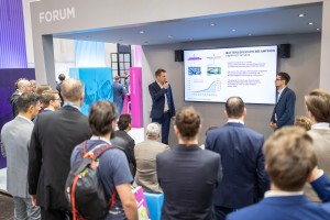 Das Foto zeigt eine Vortragssituation an einem modernen Messestand von Amprion, bei der ein größeres Publikum einem Referenten zuhört.
Vortragende und Bühne: Im Mittelpunkt unter einer grauen Überdachung mit der Aufschrift „FORUM“ steht ein Mann in einem dunkelblauen Anzug. Er hält ein Mikrofon an den Mund und spricht zu den Zuschauern. Rechts neben ihm steht ein weiterer Mann in einem blauen Sakko und blickt aufmerksam zum Sprecher.
Präsentationsinhalt: Hinter den beiden Männern ist ein großer Monitor an der Wand montiert. Die gezeigte Folie trägt die Überschrift „BATTERIESPEICHER BEI AMPRION – KNAPPHEIT IM NETZ“. Zu sehen sind Fotos von großen Batteriecontainern sowie ein Säulendiagramm mit einer ansteigenden blauen Linie, das ein deutliches Wachstum visualisiert.
Publikum: Im Bildvordergrund und links sind zahlreiche Zuschauer von hinten oder von der Seite zu sehen. Sie stehen oder sitzen konzentriert vor dem offenen Standbereich. Die Gruppe ist überwiegend männlich und in Business- oder Business-Casual-Kleidung gekleidet. Ein Mann im Vordergrund trägt einen Rucksack mit auffälligen orangefarbenen Akzenten.
Messestand: Das Design des Standes ist in Grau, Blau und Magenta gehalten. Im Hintergrund links ist eine blaue Säule mit der Aufschrift „MANAGEMENT“ und eine magenta-farbene Säule mit der Aufschrift „PROCUREMENT“ zu erkennen. Die Beleuchtung ist hell und professionell durch Deckenspots gelöst.
