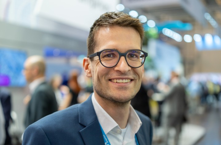 Das Porträtfoto zeigt einen jungen Mann in einer belebten, modernen Messe- oder Konferenzumgebung.
Person und Erscheinung: Der Mann blickt mit einem breiten, freundlichen Lächeln direkt in die Kamera. Er hat kurzes, dunkelbraunes Haar und trägt eine modische Brille mit dunklem, rechteckigem Rahmen.
Kleidung: Er ist professionell gekleidet mit einem dunkelblauen Sakko über einem weißen Hemd. Um den Hals trägt er ein hellblaues Schlüsselband (Lanyard), auf dem das weiße Logo von Amprion erkennbar ist.
Hintergrund und Komposition: Der Hintergrund zeigt eine unscharfe Messehalle mit hellen Lichtern, Bildschirmen und anderen Personen (Bokeh-Effekt). Dies stellt den Mann klar in den Mittelpunkt und vermittelt eine geschäftige, professionelle Atmosphäre. Das Licht ist hell und freundlich, was sein Gesicht gut ausleuchtet.
