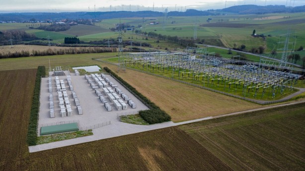 Die Luftaufnahme zeigt aus einer erhöhten Perspektive ein weitläufiges Umspannwerk (rechts) und einen direkt angrenzenden Batteriespeicher-Park (links) in einer ländlichen Umgebung.
Batteriespeicher (links): Auf einer rechteckigen, mit hellem Schotter befestigten Fläche sind über 30 große, weiße Container in zwei parallelen Doppelreihen angeordnet. Die Anlage ist von einem Schutzzaun und einer niedrigen, grünen Hecke umgeben. Am unteren Rand der Fläche befindet sich ein flaches, grünes Technikgebäude.
Umspannwerk (rechts): Direkt daneben befindet sich eine klassische Freiluft-Schaltanlage. Sie besteht aus einem dichten Netz von hohen, grauen Stahlgittermasten, Isolatorenketten und horizontal verlaufenden Stromschienen über einer Rasenfläche.
Umgebung: Die Anlagen sind in eine hügelige Landschaft mit weiten, grünen und braunen Feldern eingebettet. Im Hintergrund am Horizont sind zahlreiche Windkraftanlagen zu erkennen, was den Bezug zur Erzeugung und Speicherung erneuerbarer Energien unterstreicht. Der Himmel ist hellgrau und bedeckt.