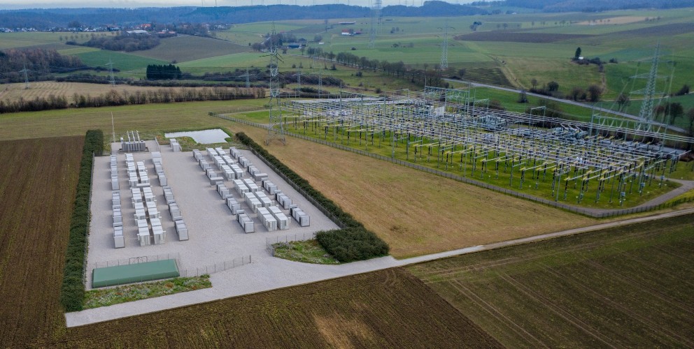 Die Luftaufnahme zeigt aus einer erhöhten Perspektive ein weitläufiges Umspannwerk (rechts) und einen direkt angrenzenden Batteriespeicher-Park (links) in einer ländlichen Umgebung.
Batteriespeicher (links): Auf einer rechteckigen, mit hellem Schotter befestigten Fläche sind über 30 große, weiße Container in zwei parallelen Doppelreihen angeordnet. Die Anlage ist von einem Schutzzaun und einer niedrigen, grünen Hecke umgeben. Am unteren Rand der Fläche befindet sich ein flaches, grünes Technikgebäude.
Umspannwerk (rechts): Direkt daneben befindet sich eine klassische Freiluft-Schaltanlage. Sie besteht aus einem dichten Netz von hohen, grauen Stahlgittermasten, Isolatorenketten und horizontal verlaufenden Stromschienen über einer Rasenfläche.
Umgebung: Die Anlagen sind in eine hügelige Landschaft mit weiten, grünen und braunen Feldern eingebettet. Im Hintergrund am Horizont sind zahlreiche Windkraftanlagen zu erkennen, was den Bezug zur Erzeugung und Speicherung erneuerbarer Energien unterstreicht. Der Himmel ist hellgrau und bedeckt.