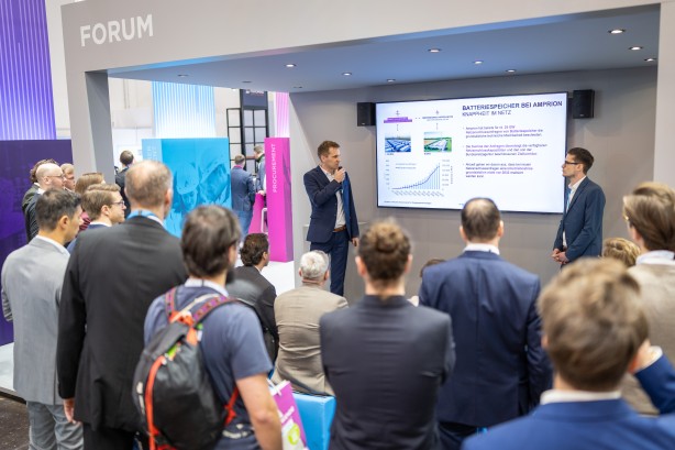 Das Foto zeigt eine Vortragssituation an einem modernen Messestand von Amprion, bei der ein größeres Publikum einem Referenten zuhört.
Vortragende und Bühne: Im Mittelpunkt unter einer grauen Überdachung mit der Aufschrift „FORUM“ steht ein Mann in einem dunkelblauen Anzug. Er hält ein Mikrofon an den Mund und spricht zu den Zuschauern. Rechts neben ihm steht ein weiterer Mann in einem blauen Sakko und blickt aufmerksam zum Sprecher.
Präsentationsinhalt: Hinter den beiden Männern ist ein großer Monitor an der Wand montiert. Die gezeigte Folie trägt die Überschrift „BATTERIESPEICHER BEI AMPRION – KNAPPHEIT IM NETZ“. Zu sehen sind Fotos von großen Batteriecontainern sowie ein Säulendiagramm mit einer ansteigenden blauen Linie, das ein deutliches Wachstum visualisiert.
Publikum: Im Bildvordergrund und links sind zahlreiche Zuschauer von hinten oder von der Seite zu sehen. Sie stehen oder sitzen konzentriert vor dem offenen Standbereich. Die Gruppe ist überwiegend männlich und in Business- oder Business-Casual-Kleidung gekleidet. Ein Mann im Vordergrund trägt einen Rucksack mit auffälligen orangefarbenen Akzenten.
Messestand: Das Design des Standes ist in Grau, Blau und Magenta gehalten. Im Hintergrund links ist eine blaue Säule mit der Aufschrift „MANAGEMENT“ und eine magenta-farbene Säule mit der Aufschrift „PROCUREMENT“ zu erkennen. Die Beleuchtung ist hell und professionell durch Deckenspots gelöst.
