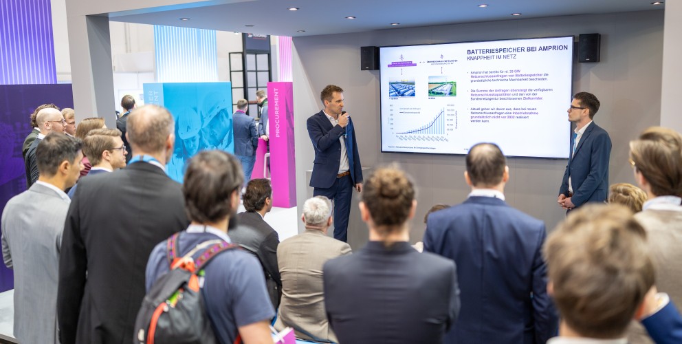 Das Foto zeigt eine Vortragssituation an einem modernen Messestand von Amprion, bei der ein größeres Publikum einem Referenten zuhört.
Vortragende und Bühne: Im Mittelpunkt unter einer grauen Überdachung mit der Aufschrift „FORUM“ steht ein Mann in einem dunkelblauen Anzug. Er hält ein Mikrofon an den Mund und spricht zu den Zuschauern. Rechts neben ihm steht ein weiterer Mann in einem blauen Sakko und blickt aufmerksam zum Sprecher.
Präsentationsinhalt: Hinter den beiden Männern ist ein großer Monitor an der Wand montiert. Die gezeigte Folie trägt die Überschrift „BATTERIESPEICHER BEI AMPRION – KNAPPHEIT IM NETZ“. Zu sehen sind Fotos von großen Batteriecontainern sowie ein Säulendiagramm mit einer ansteigenden blauen Linie, das ein deutliches Wachstum visualisiert.
Publikum: Im Bildvordergrund und links sind zahlreiche Zuschauer von hinten oder von der Seite zu sehen. Sie stehen oder sitzen konzentriert vor dem offenen Standbereich. Die Gruppe ist überwiegend männlich und in Business- oder Business-Casual-Kleidung gekleidet. Ein Mann im Vordergrund trägt einen Rucksack mit auffälligen orangefarbenen Akzenten.
Messestand: Das Design des Standes ist in Grau, Blau und Magenta gehalten. Im Hintergrund links ist eine blaue Säule mit der Aufschrift „MANAGEMENT“ und eine magenta-farbene Säule mit der Aufschrift „PROCUREMENT“ zu erkennen. Die Beleuchtung ist hell und professionell durch Deckenspots gelöst.
